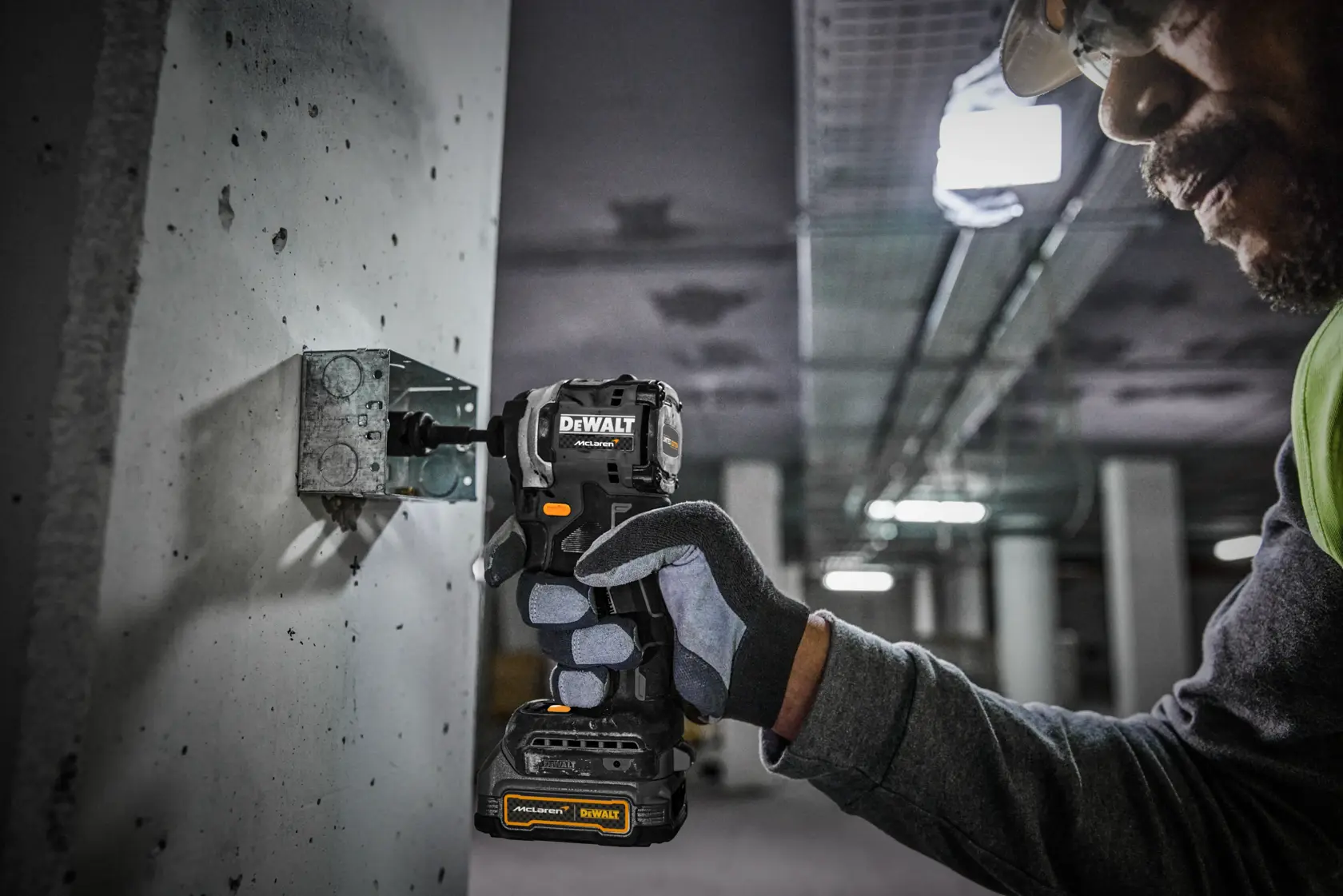 A person wearing gloves and safety gear uses a DEWALT power tool to fasten a metal bracket to a concrete wall inside an industrial building.