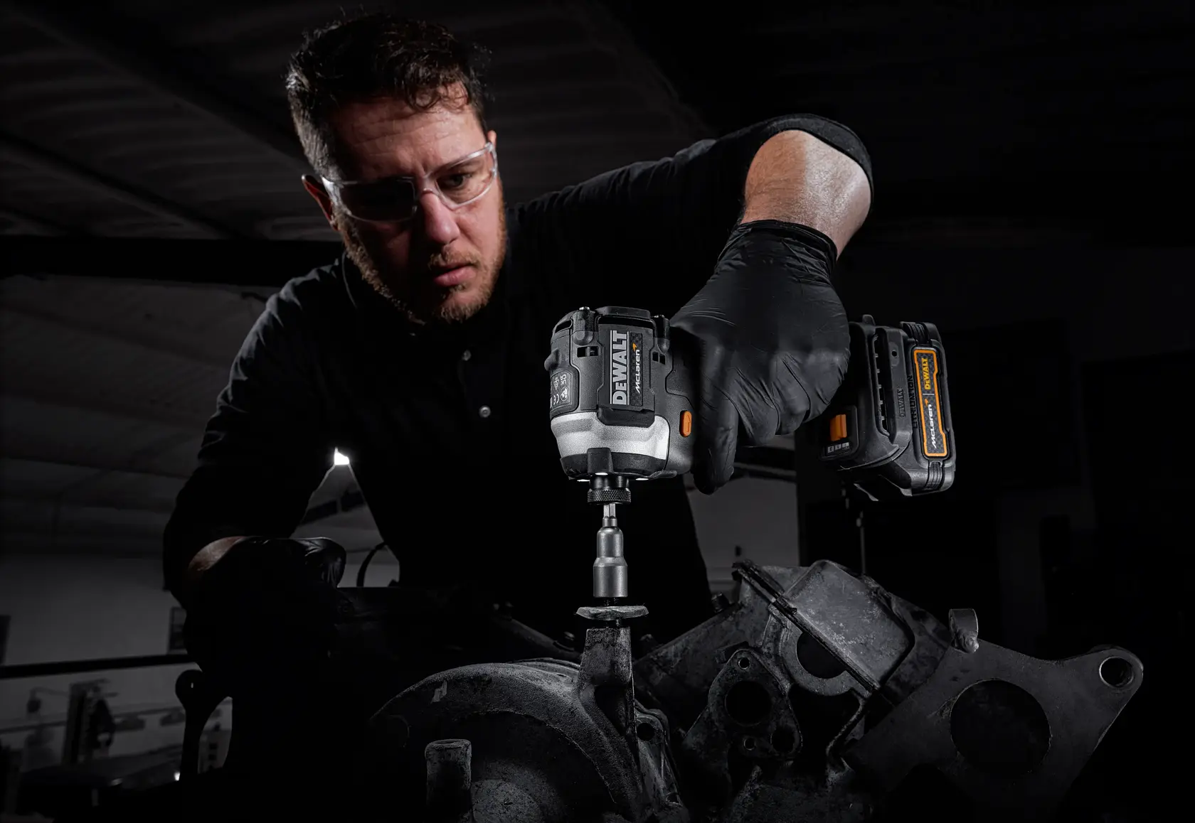 A person with a blurred face uses a DEWALT cordless tool, model DCF85ME2GT, to work on mechanical equipment. The person is wearing black gloves and is in a workshop setting.