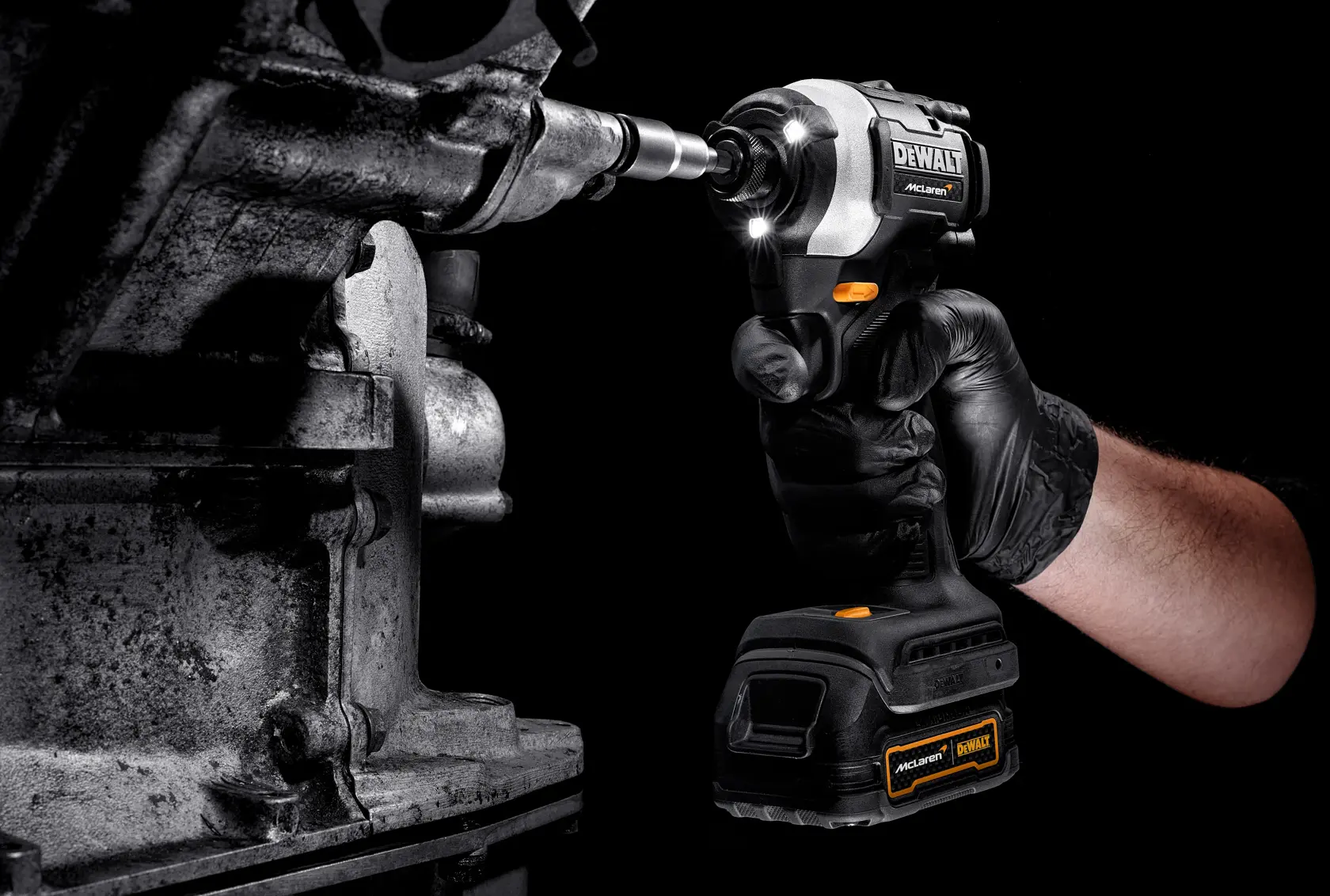 A person wearing a black glove uses a DEWALT cordless impact driver to tighten or loosen a bolt on a metallic machine part. The tool is equipped with LED lights illuminating the work area.