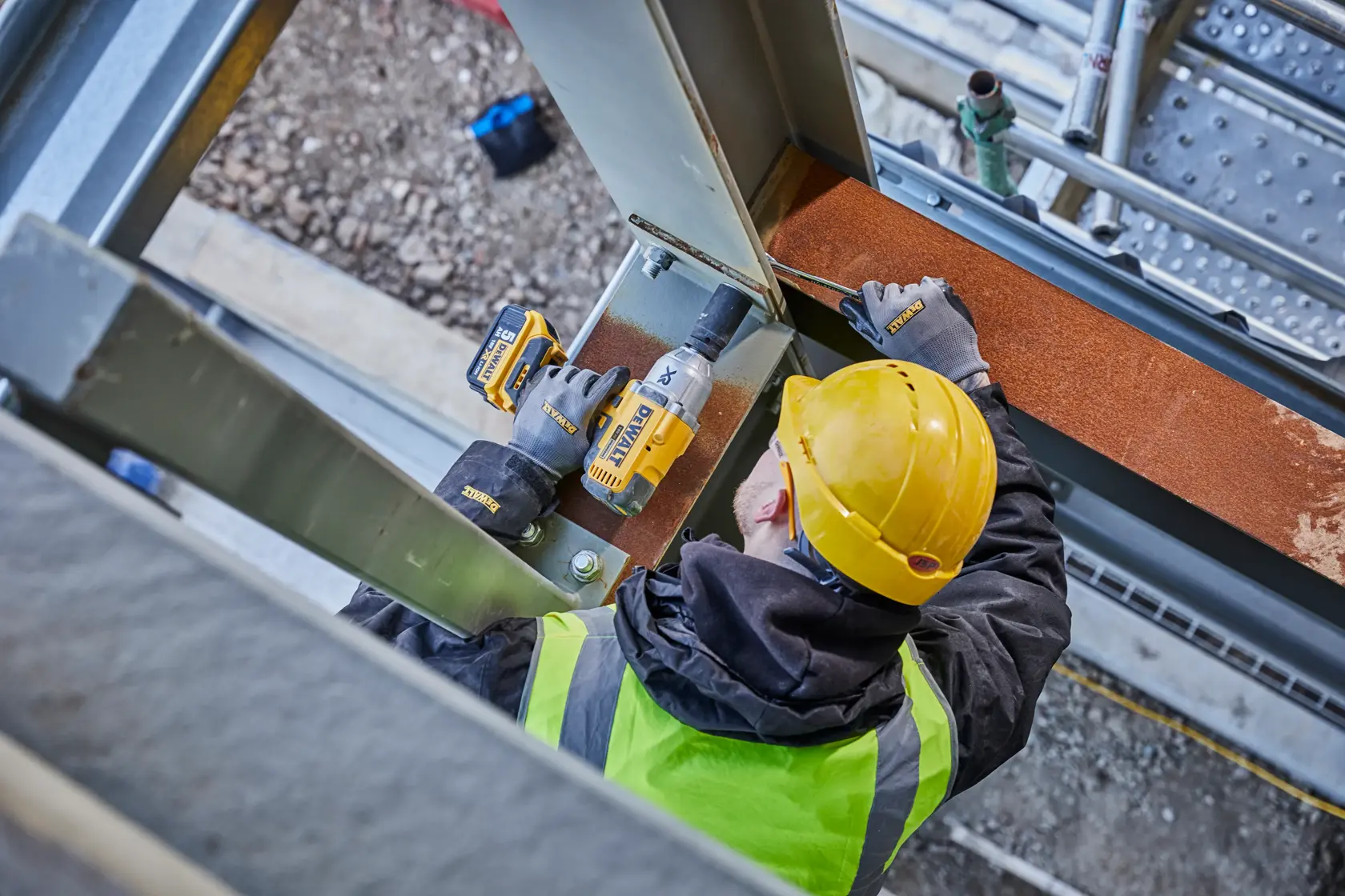 A person wearing a yellow hard hat and high-visibility vest uses a DEWALT cordless impact wrench to fasten bolts on a steel beam at a construction site.