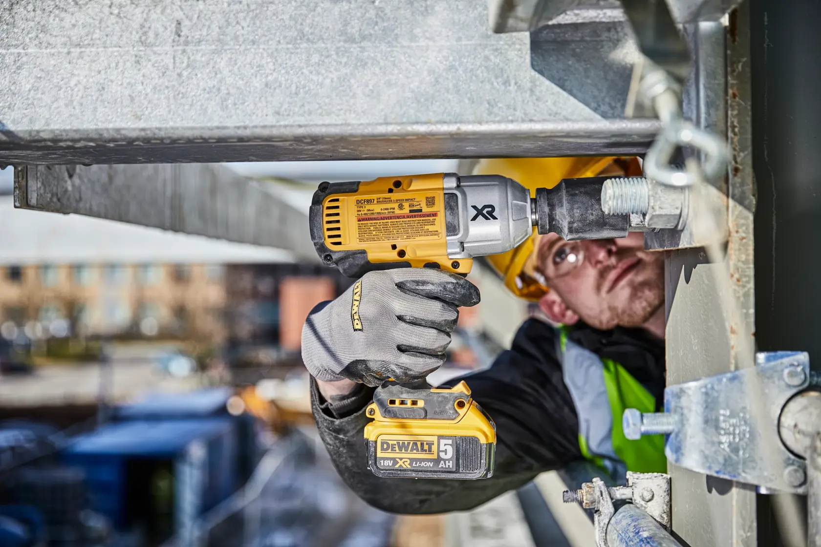 A person wearing protective gloves and a helmet uses a DEWALT cordless impact wrench (model DCF897N) to fasten a large bolt on a steel structure at a construction site.