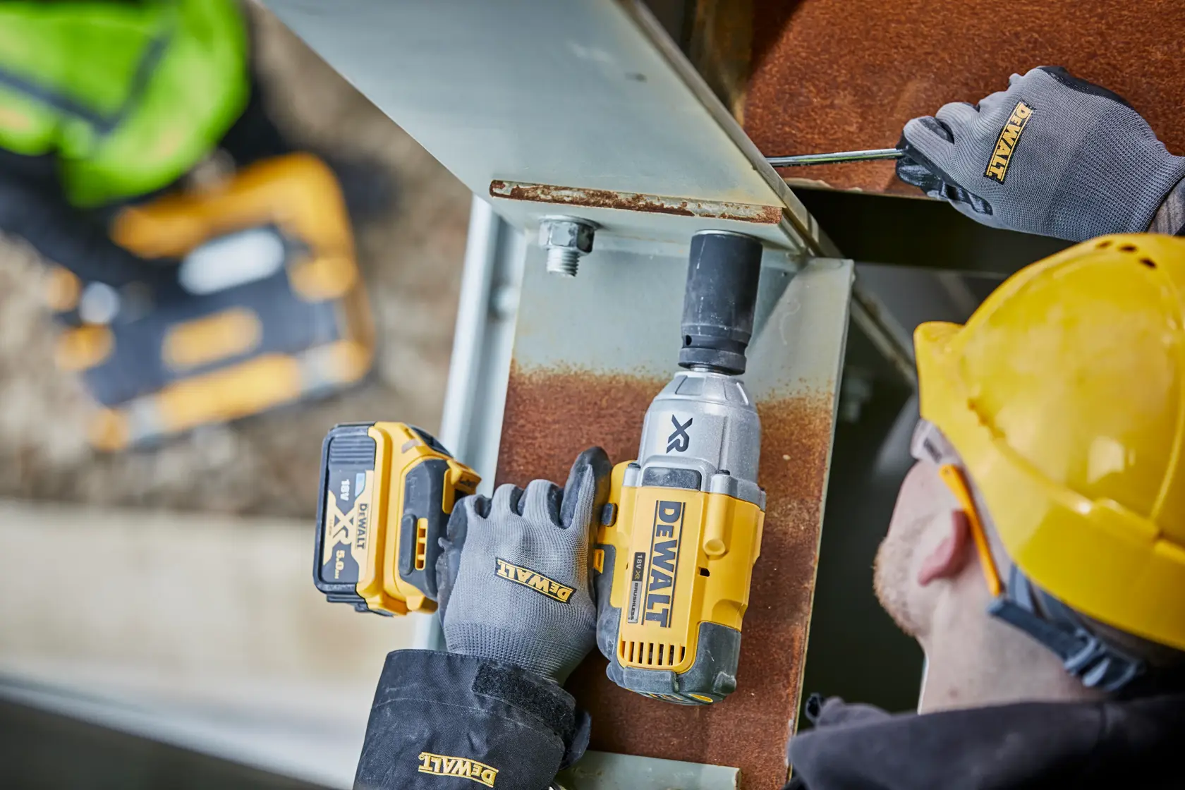 A worker wearing a yellow hard hat and DEWALT gloves uses a DEWALT cordless impact wrench (model DCF897N) on a steel beam at a construction site.