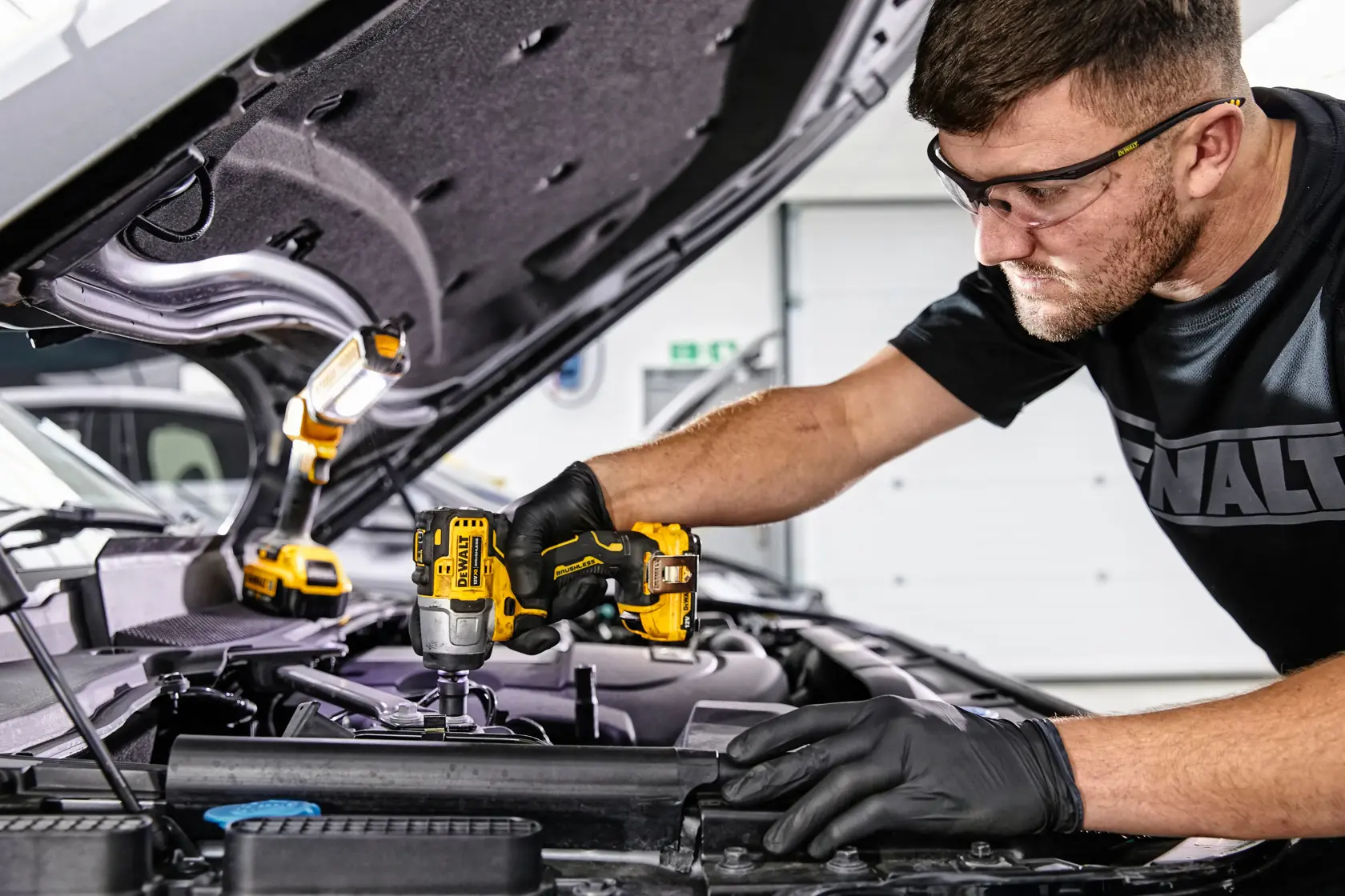 A person wearing black gloves uses a yellow DEWALT cordless impact wrench (SKU: DCF902D2) to work on the engine of a car. An additional DEWALT work light is positioned under the raised hood for illumination.