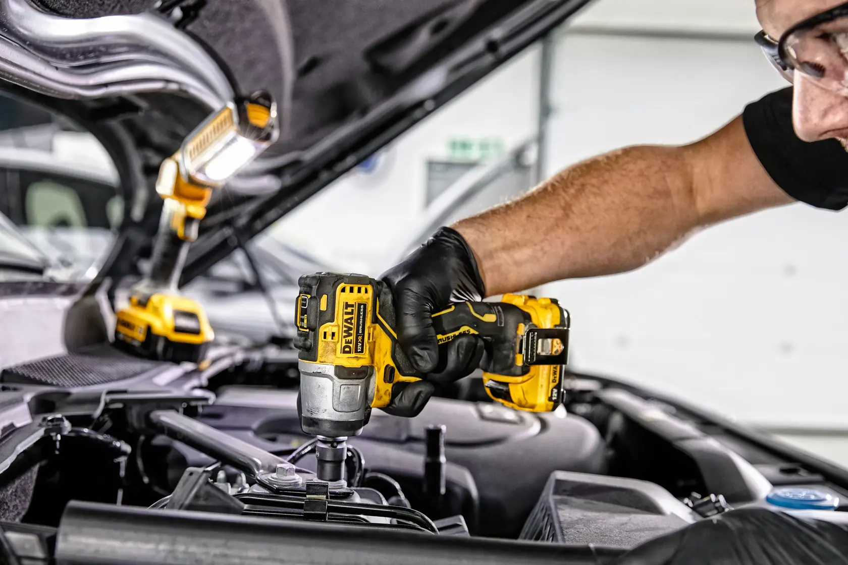 A person wearing gloves uses a DEWALT DCF902D2 cordless impact wrench to work on a car engine, with a DEWALT work light illuminating the area.