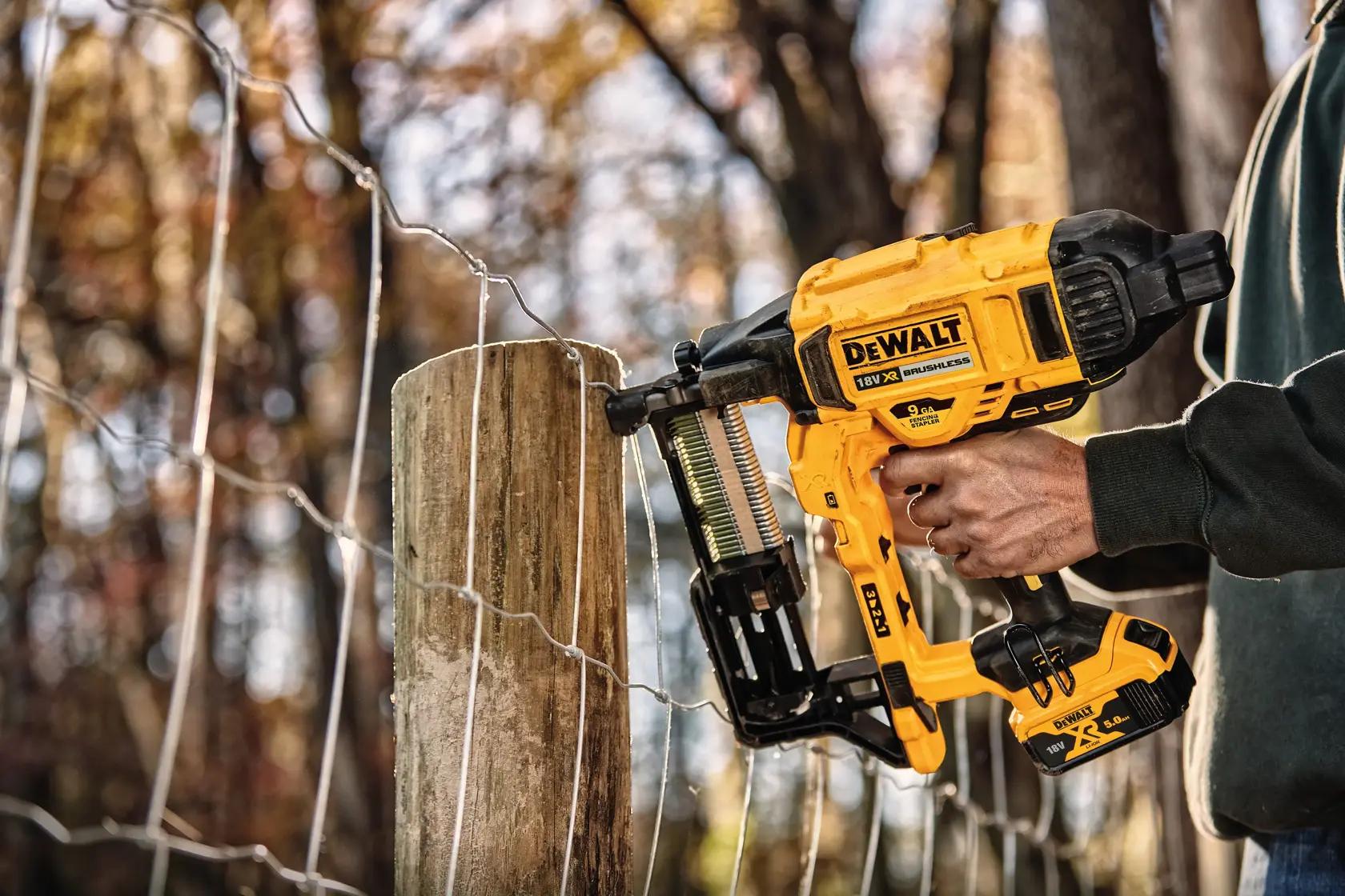 A person uses a yellow DEWALT cordless fencing stapler to attach wire fencing to a wooden post outdoors.