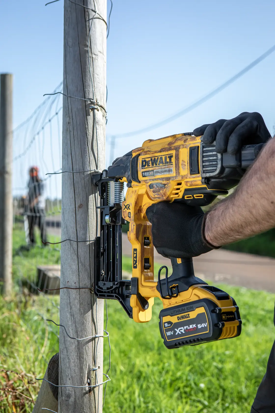 DEWALT cordless fencing stapler is used to fasten wire to a wooden post outdoors.