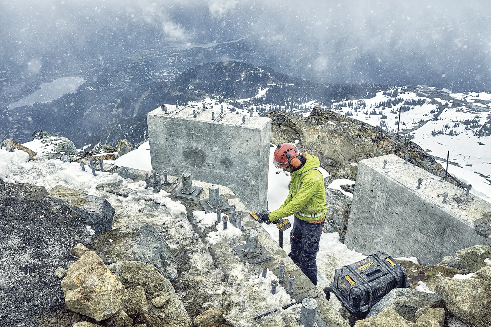 Worker uses DEWALT tool outdoors in snowy mountain area near concrete blocks, with DEWALT case on the ground.
