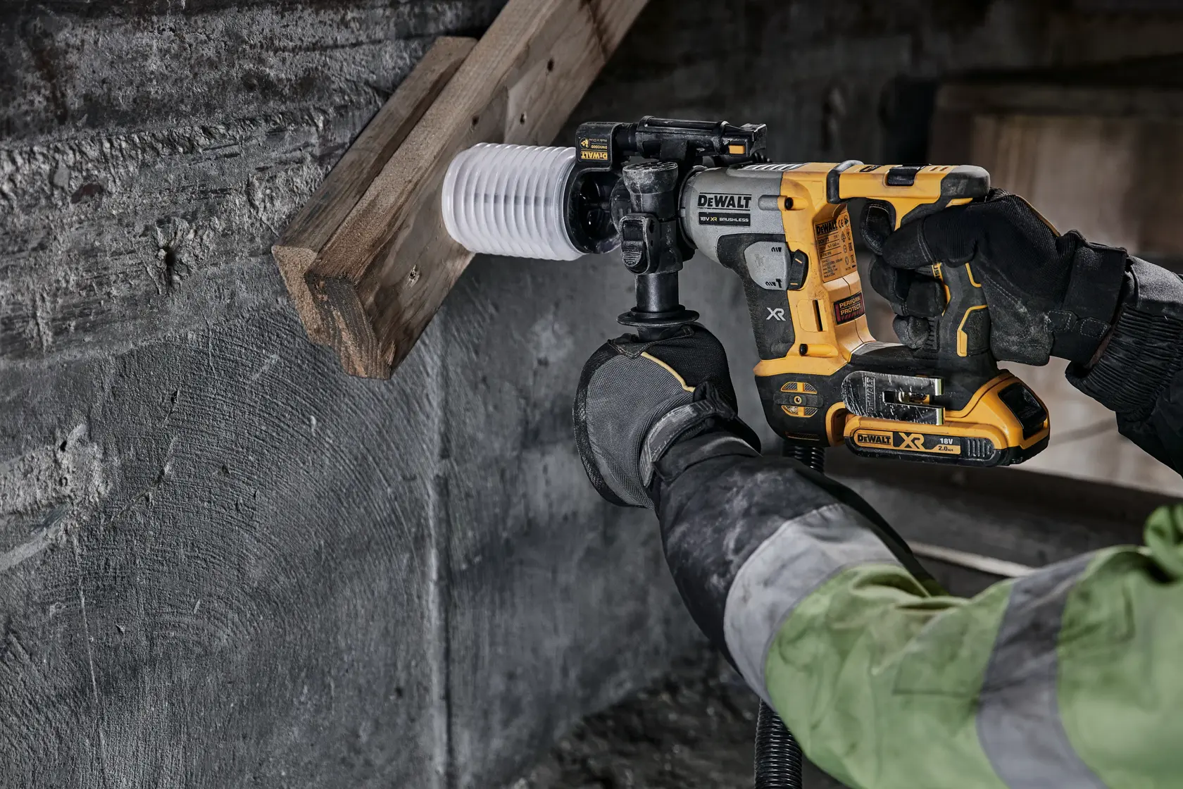 DEWALT cordless hammer drill in use, drilling a hole through wood and concrete by a worker wearing gloves.