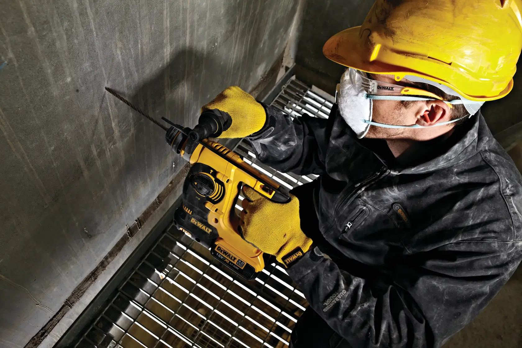 Worker wearing safety gear uses a DEWALT rotary hammer drill to bore into a concrete wall at an industrial site.