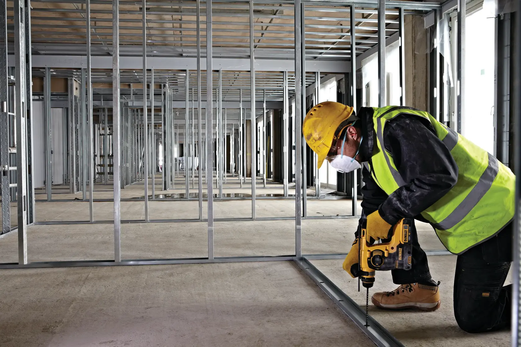 Worker with yellow drill installs metal frame inside a building under construction, wearing safety gear.