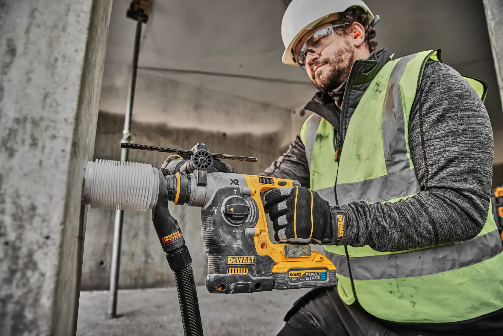 A construction worker wearing a safety helmet and high-visibility vest uses a DEWALT DCH273H2T cordless rotary hammer drill to drill into a concrete column. The worker is also wearing gloves branded with DEWALT.