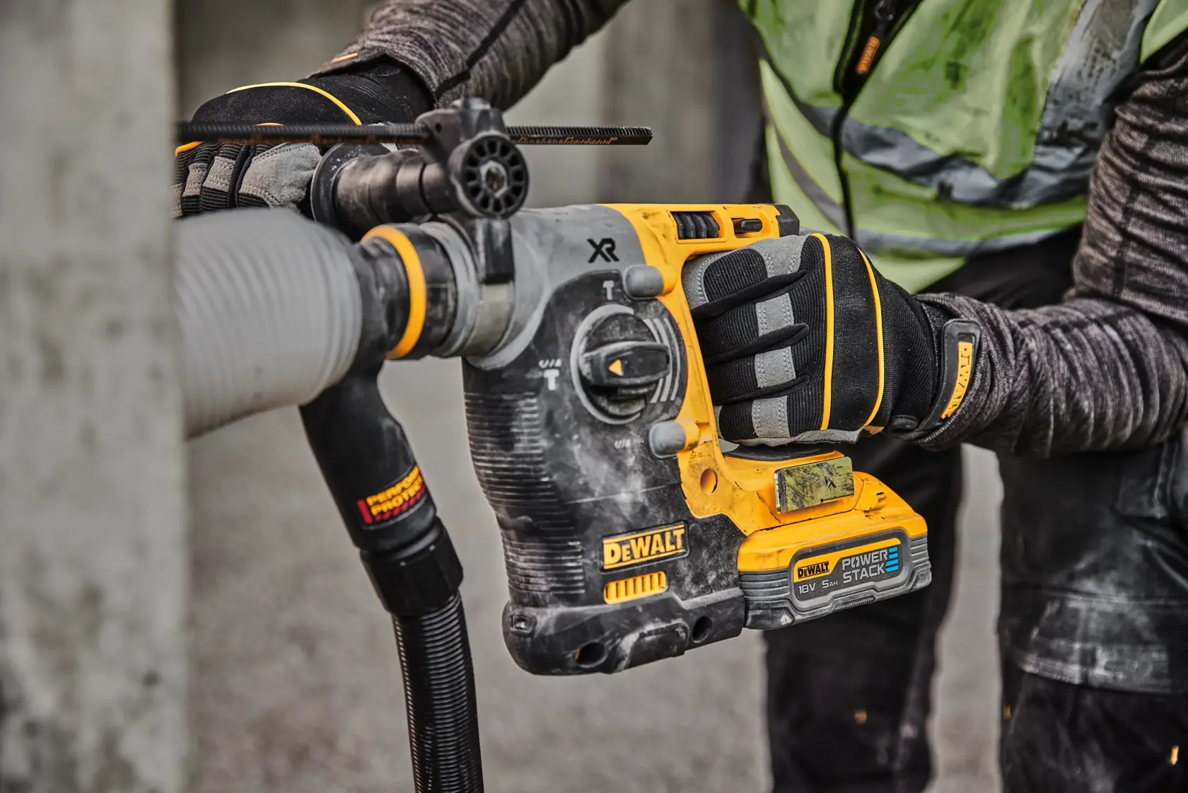 A close-up of a worker using a DEWALT cordless rotary hammer drill with a POWERSTACK battery to drill into concrete, wearing protective gloves and clothing.