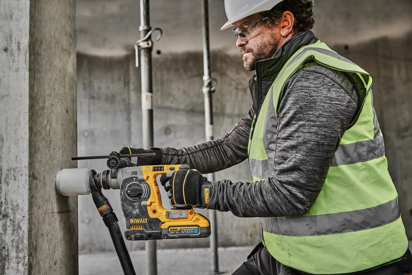 A person wearing a safety vest and gloves is using a DEWALT DCH273H2T cordless rotary hammer drill to drill into a concrete pillar at a construction site. The user's face is blurred for privacy.