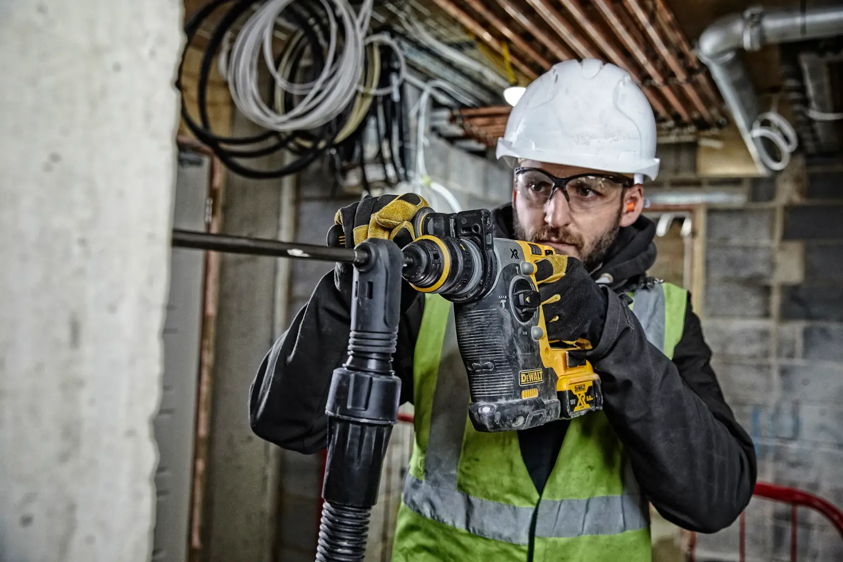 A construction worker wearing a hard hat and safety vest is operating a DEWALT DCH274P2 cordless rotary hammer drill inside a building under construction. The worker is using the tool to drill into a concrete surface.