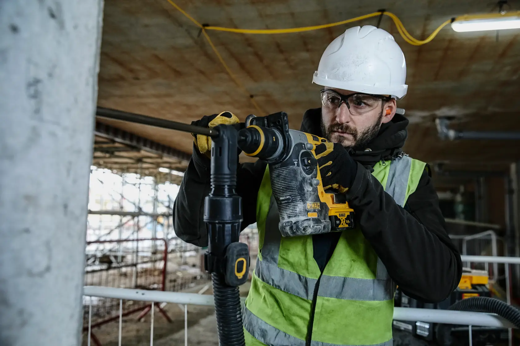 A person wearing a safety helmet and high-visibility vest operates a DEWALT DCH274P2 rotary hammer drill at a construction site.