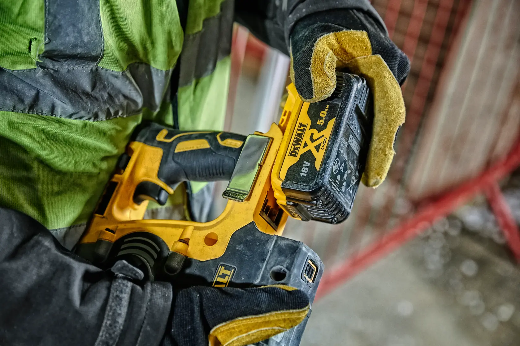 A person wearing gloves and a green work jacket is holding a DEWALT cordless power drill with an 18V XR 5.0Ah battery attached.