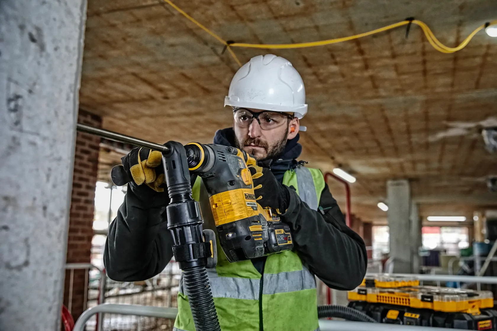 Worker using a DEWALT DCH323T2 rotary hammer drill on concrete at a construction site, wearing safety gear.