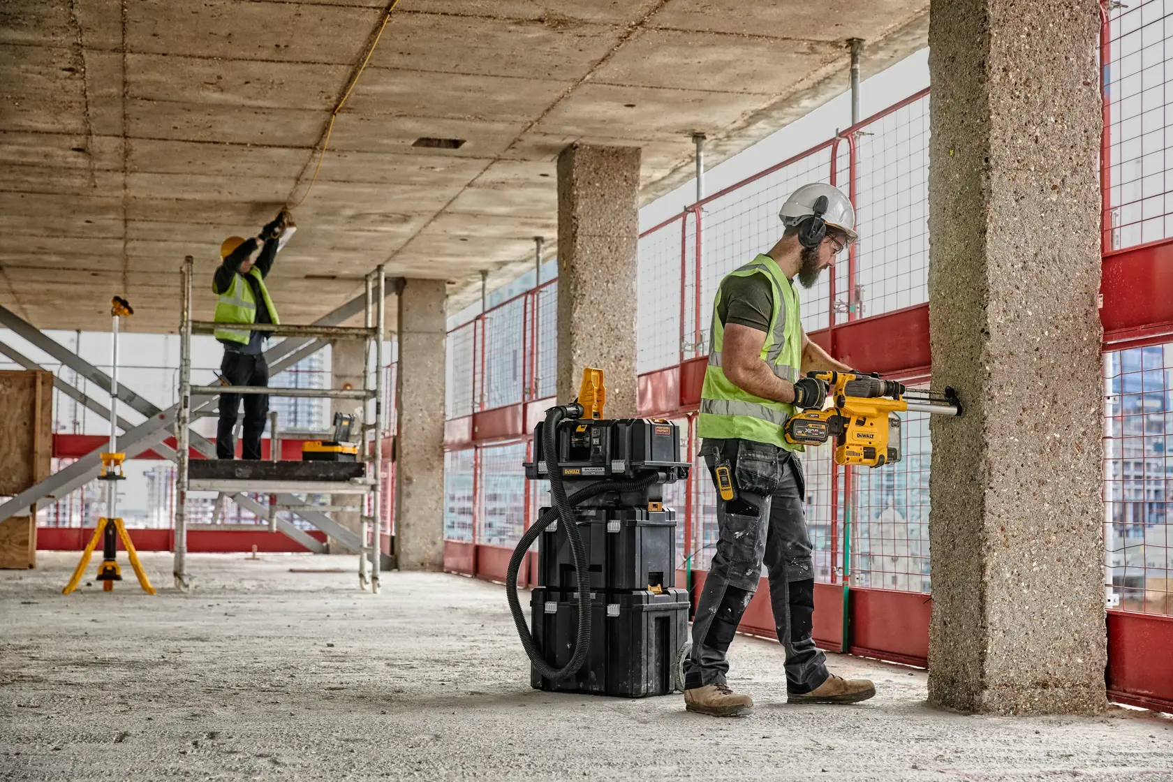 Two construction workers are operating DEWALT power tools at a building site. One worker is drilling into a concrete pillar, wearing a hard hat, safety vest, and protective gear, while the other worker is standing on scaffolding measuring the ceiling. DEWALT toolboxes and equipment are visible nearby.