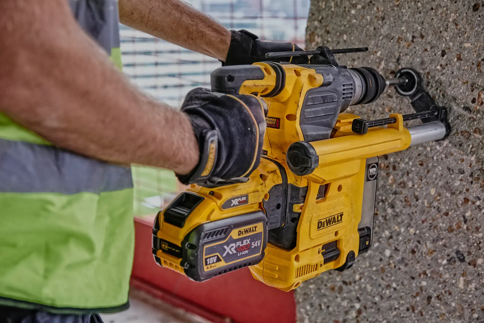 A person wearing work gloves and a safety vest is using a DEWALT cordless rotary hammer drill to bore into a concrete wall.