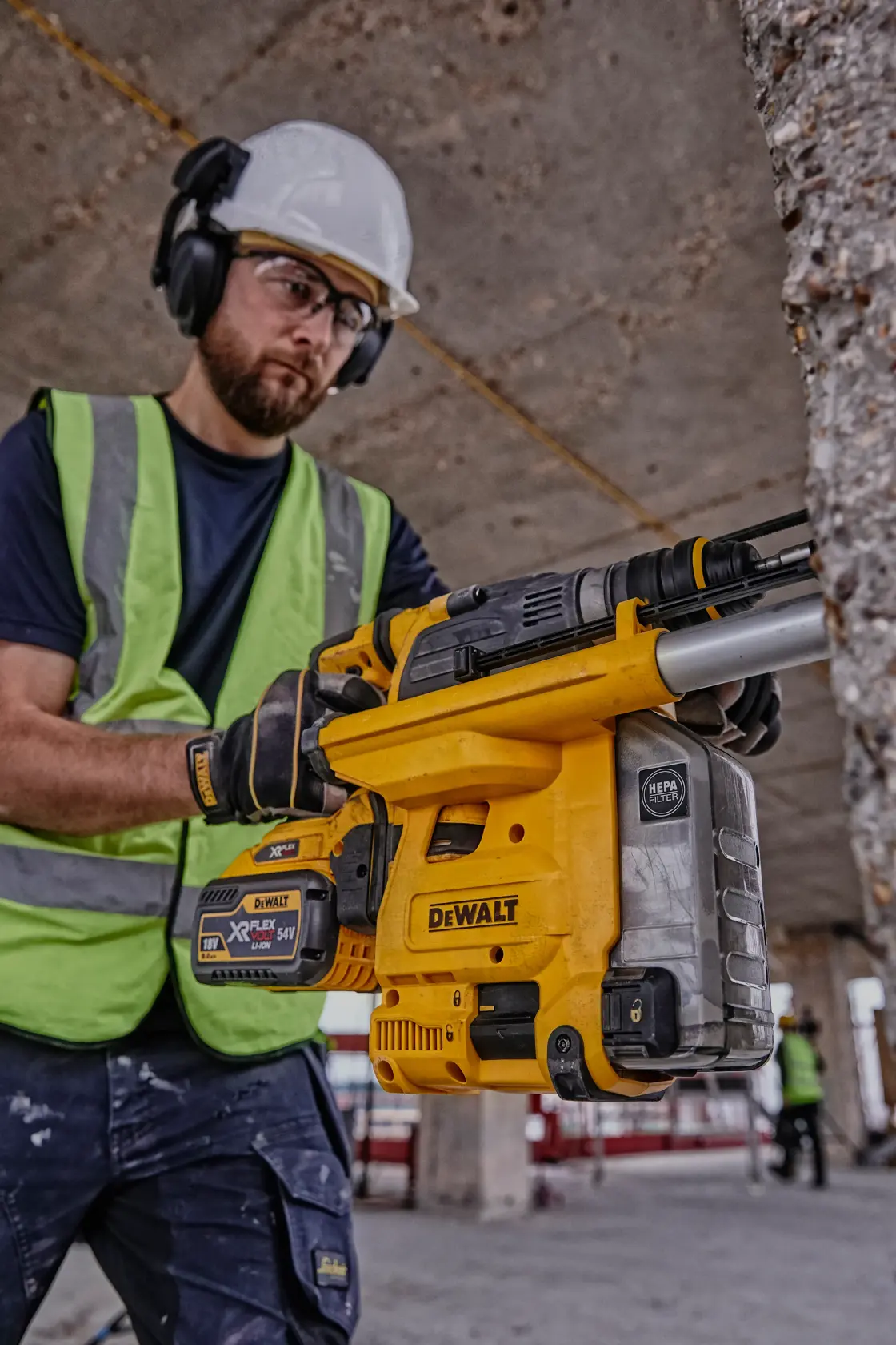 Worker holding a DEWALT DCH334X2 tool at a construction site, wearing safety gear and a green vest.