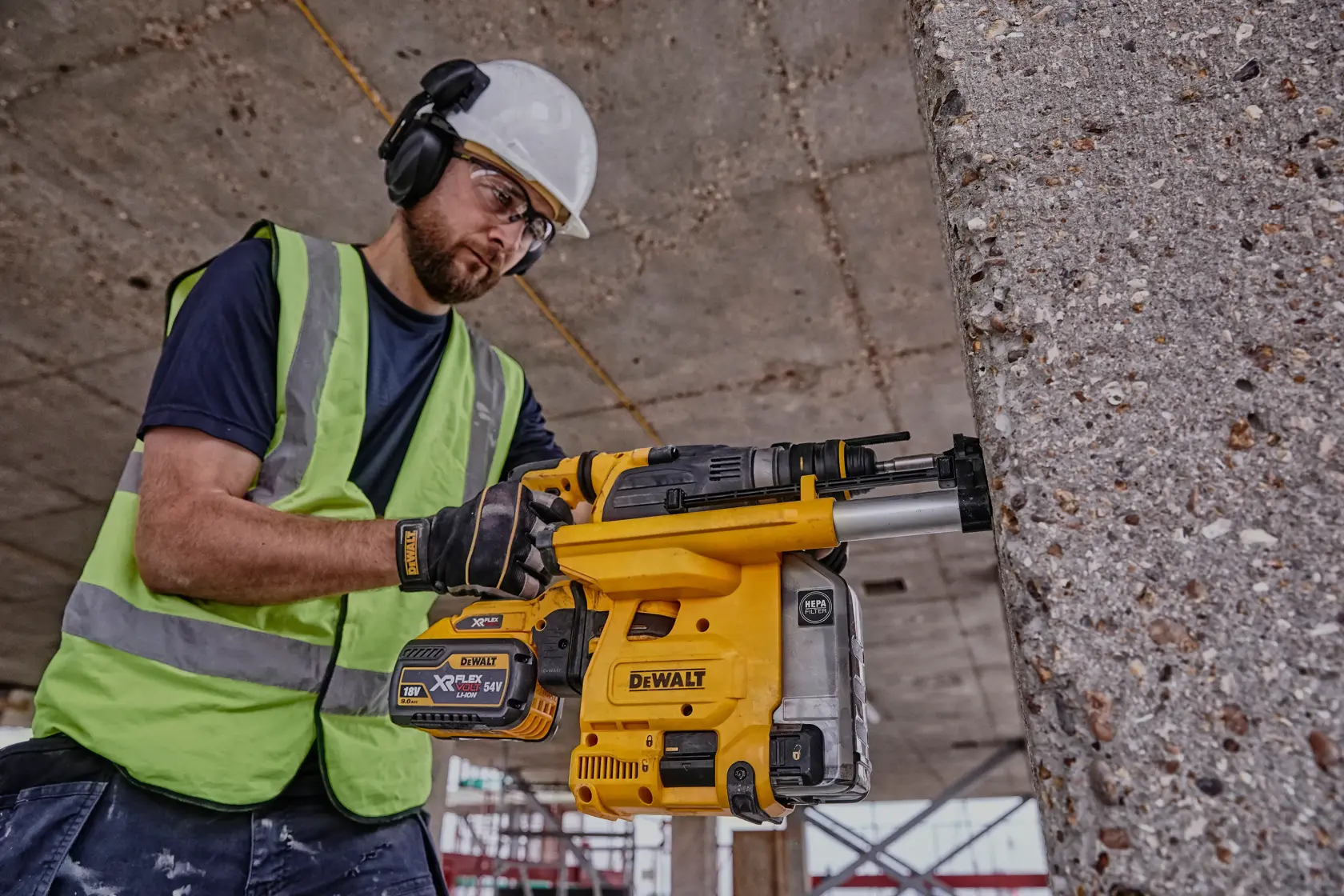 A person wearing safety gear and a high-visibility vest operates a yellow DEWALT DCH334X2 rotary hammer drill against a concrete wall at a construction site.
