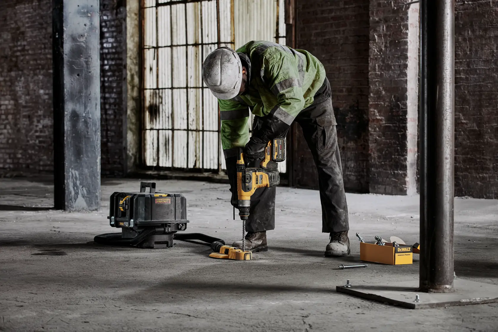 Worker drills concrete floor using DEWALT power tool; toolbox and equipment visible nearby in industrial setting.