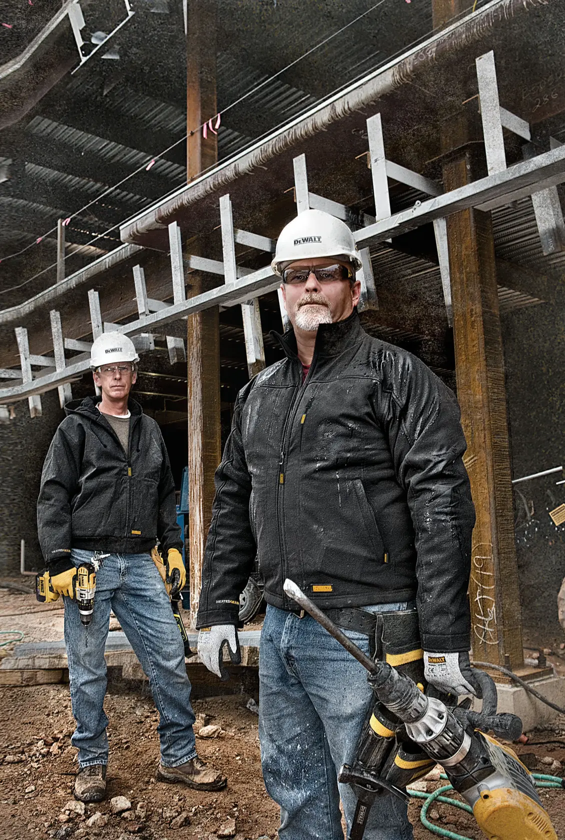 Two workers wearing DEWALT-branded safety helmets and black jackets stand at a construction site. One worker holds a yellow power tool. The workers' faces are blurred. The site is partially built with metal and wood structures visible.