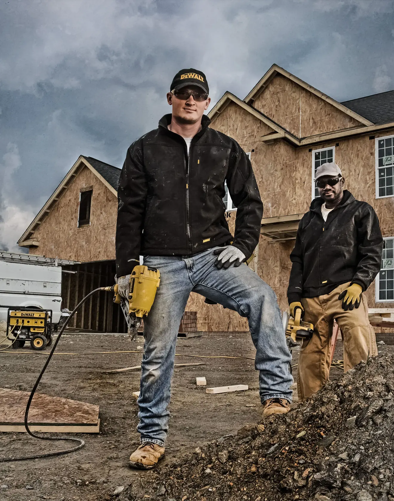 Two workers wearing black jackets and gloves stand in front of a residential construction site. One worker holds a power tool and wears a DEWALT-branded cap, while the other stands behind holding another tool. DEWALT products and an unfinished house are visible in the background.