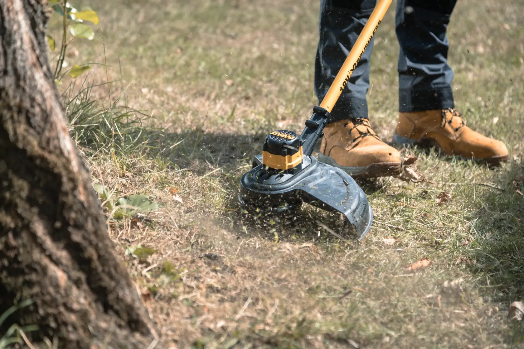 Person using a DEWALT cordless grass trimmer outdoors near a tree, cutting grass while wearing work boots.