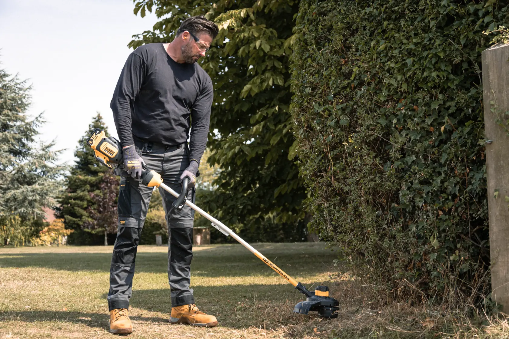 Person using DEWALT grass trimmer outdoors, trimming grass near hedge on a sunny day.