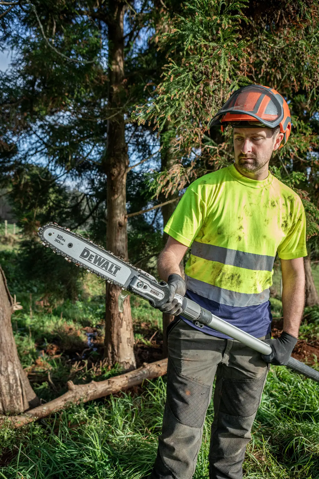 Person outdoors holding DEWALT pole saw DCMPS640N, wearing safety gear and work clothes, with trees in background.