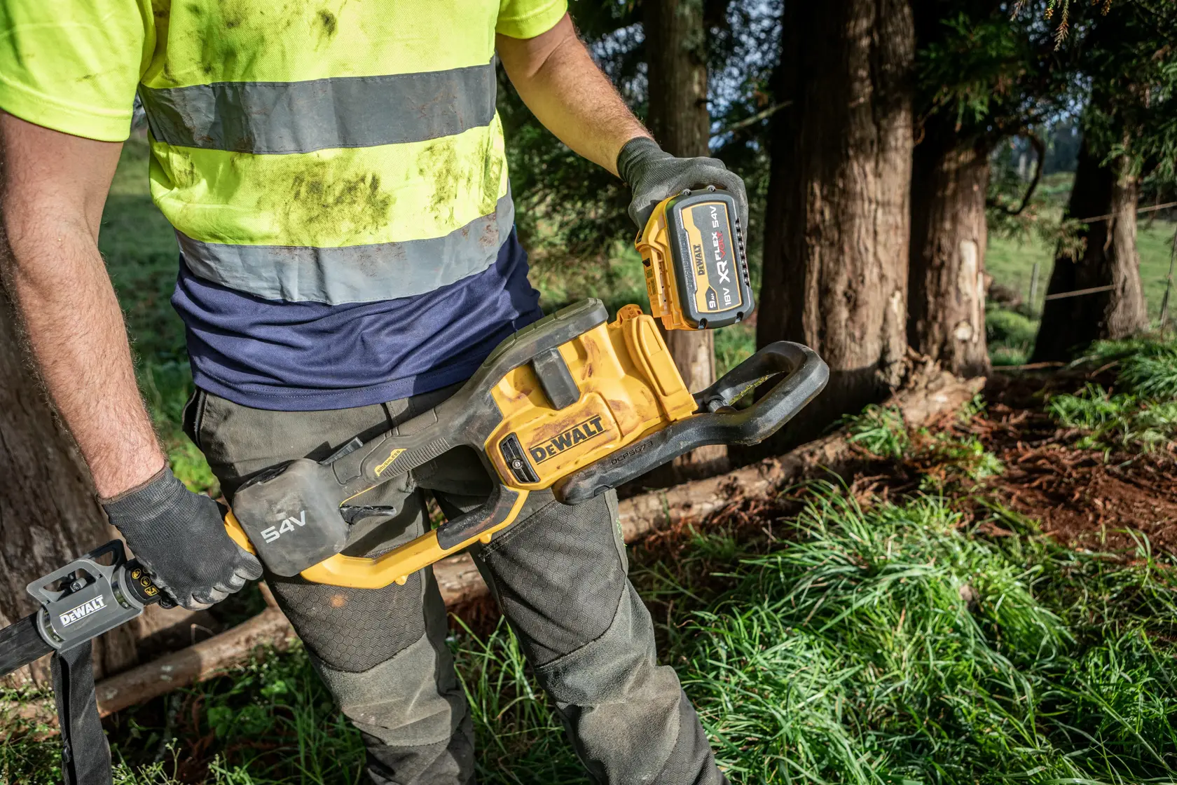 Person holding a DEWALT cordless chainsaw outdoors, wearing work gloves and a high-visibility shirt.