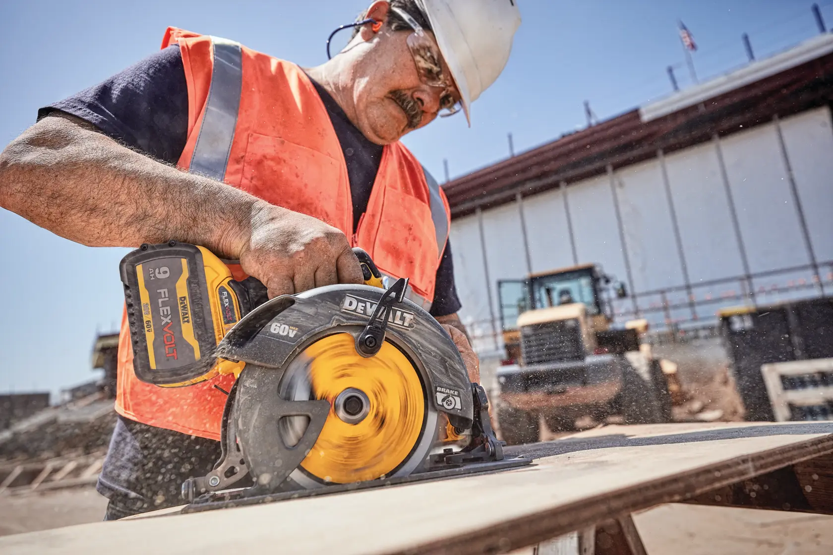 A construction worker wearing a safety vest and helmet uses a DEWALT DCS578X2 60V FLEXVOLT cordless circular saw to cut a wooden board at an outdoor construction site. The saw is in use, producing visible sawdust.