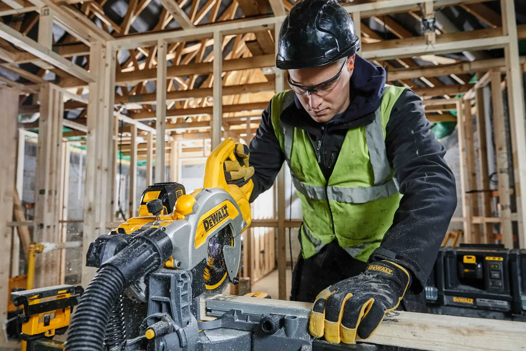 Worker uses DEWALT DCS777T2 saw to cut wood at a construction site, wearing safety gear and gloves.