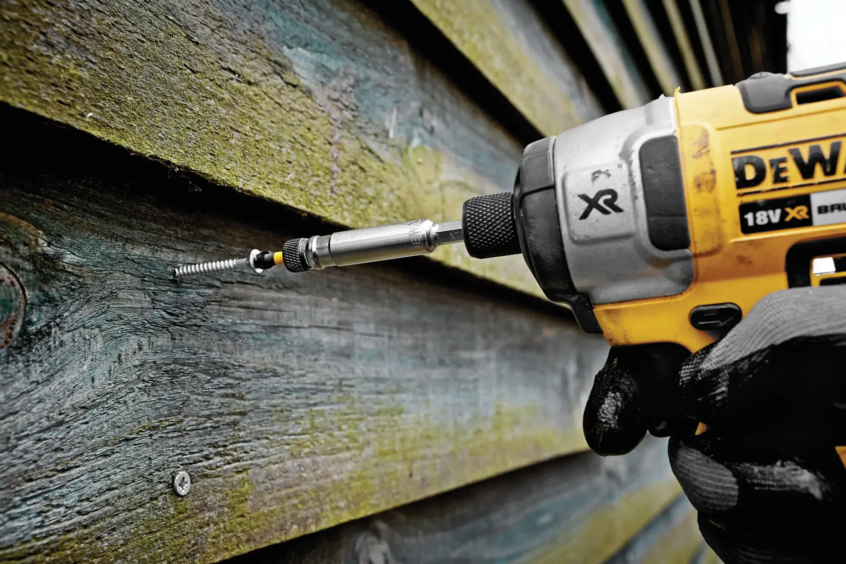 A DEWALT driver tool is screwing a fastener into wooden planks outdoors, held by a gloved hand.
