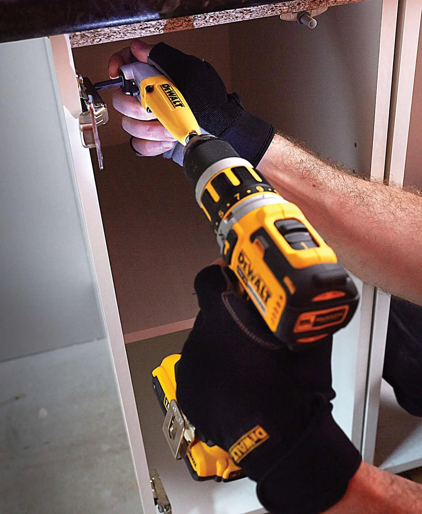 Person using a DEWALT drill inside a cabinet, wearing black gloves, assembling or fixing hinge hardware.