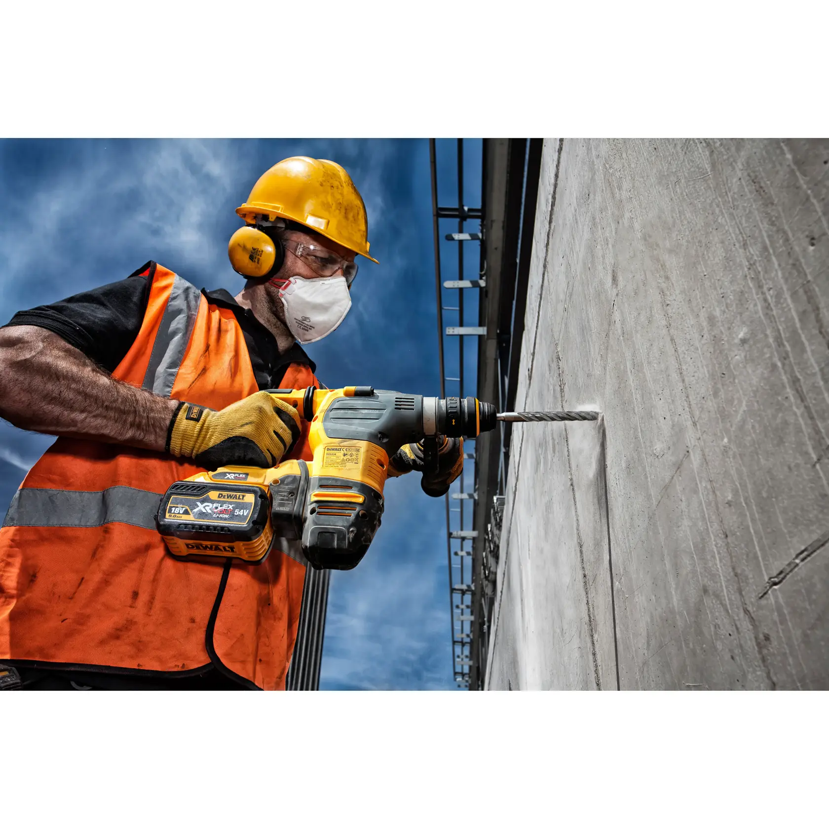 Un ouvrier du bâtiment portant un casque jaune, un gilet de sécurité orange et des gants utilise un perforateur sans fil DEWALT XR avec un foret DT9529 pour percer un mur en béton à l'extérieur.