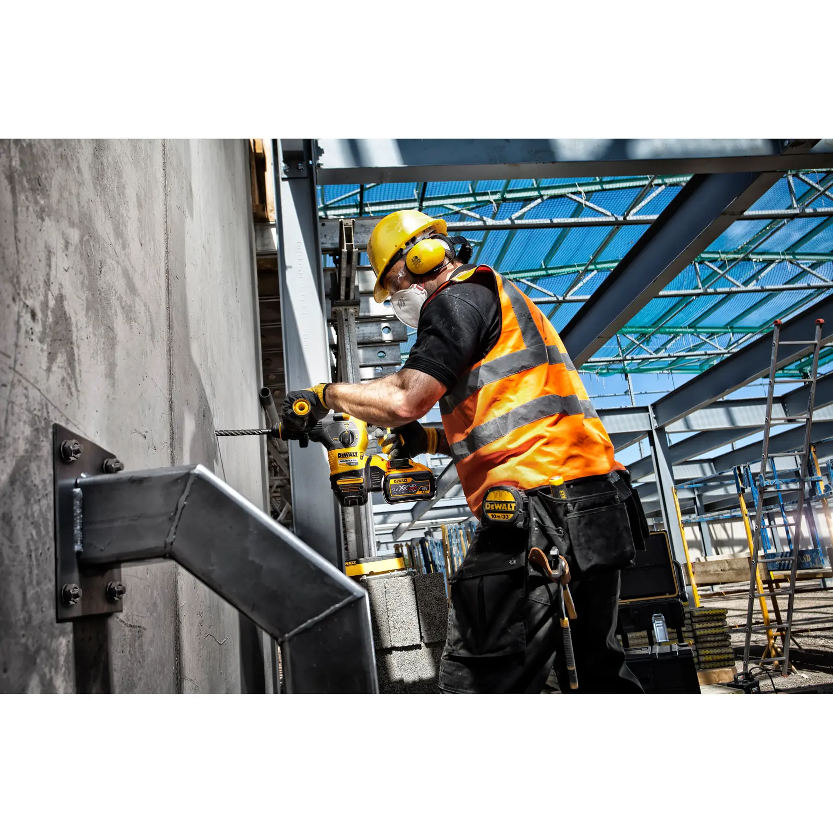 Un ouvrier du bâtiment portant un casque jaune, une protection auditive, des lunettes de sécurité, un gilet réfléchissant orange et un masque utilise une perceuse DEWALT pour percer un mur en béton sur un chantier industriel.