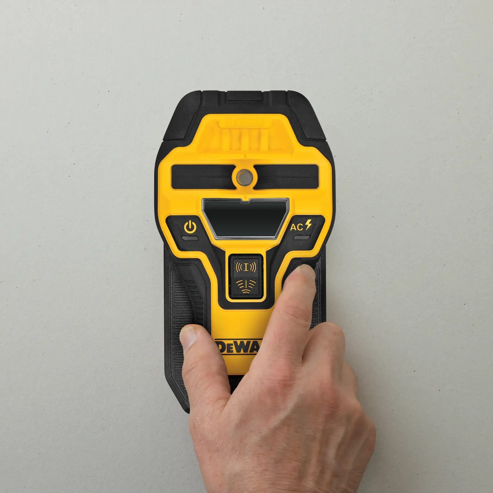 A hand is holding a yellow and black DEWALT electronic device, likely a wall scanner or stud finder, positioned against a light grey surface.