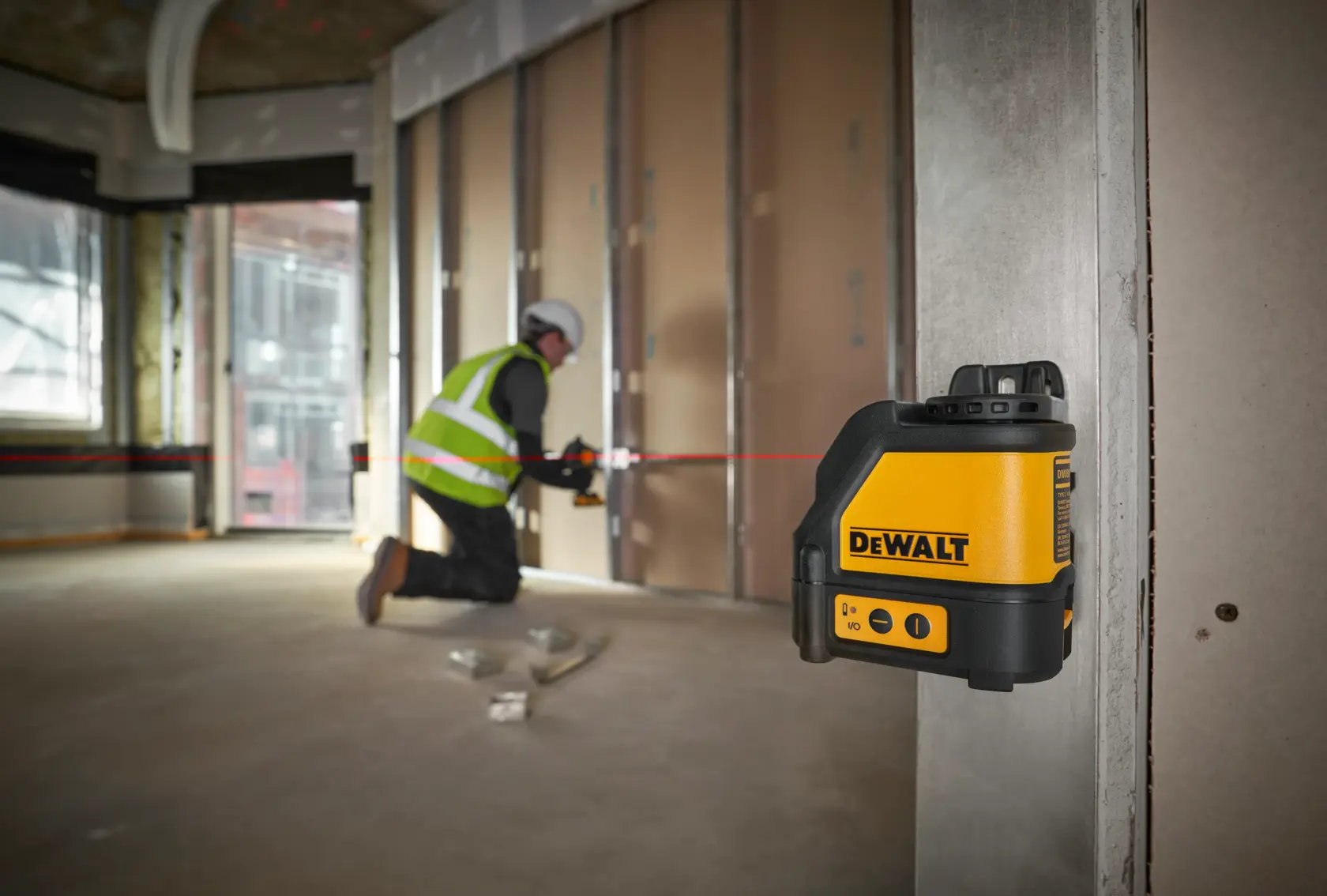 A person wearing a hard hat, safety vest, and gloves is kneeling on the floor of a construction site, using a DEWALT DW088K laser level. The device is projecting a visible red laser line for alignment. The surroundings show an unfinished building with concrete columns and open windows.