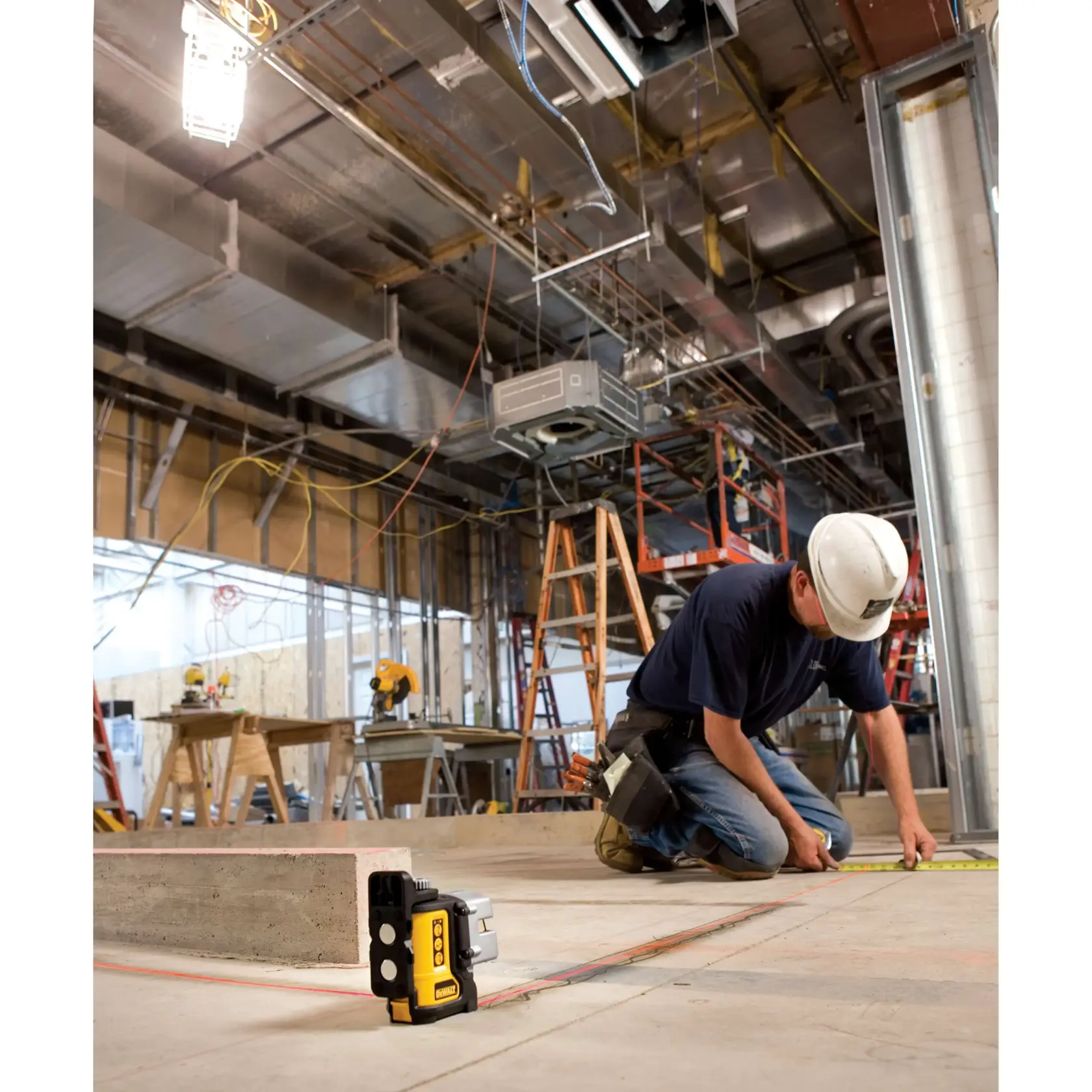 Deux ouvriers du bâtiment assemblent des structures métalliques sur un chantier intérieur. Un ouvrier est debout sur une échelle verte, tandis que l'autre est agenouillé sur un échafaudage jaune. Un niveau laser DEWALT DW089K est posé au sol, projetant des lignes laser rouges pour aider à l’alignement. L’espace de travail est rempli d’équipements de construction, de conduits et d’échafaudages.