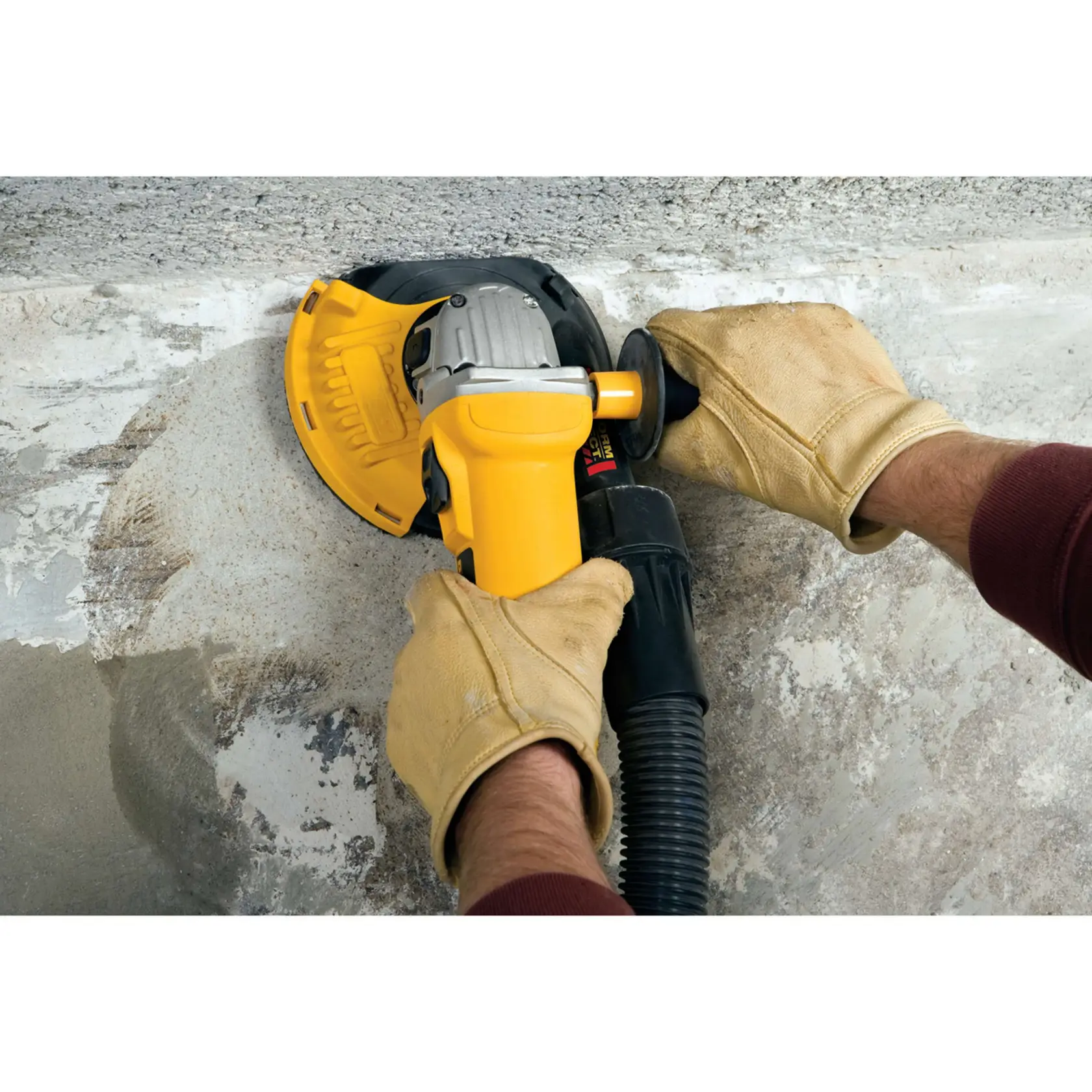 Un ouvrier de construction portant un casque jaune, des gants et un masque facial utilise un outil de ponçage de surface DEWALT DWE46150 sur un sol en béton, avec un aspirateur extracteur de poussière jaune DEWALT en arrière-plan. La scène se déroule dans un environnement industriel ou de construction.