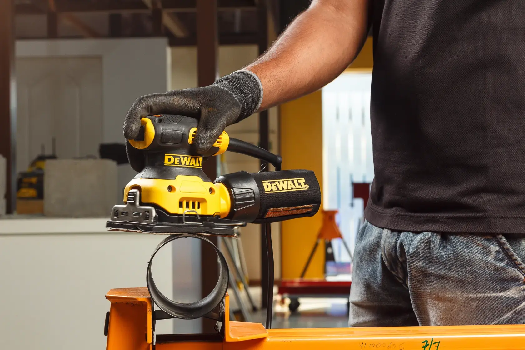 A close-up of a person wearing gloves using a yellow DEWALT electric sander to smooth the surface of a wooden plank.