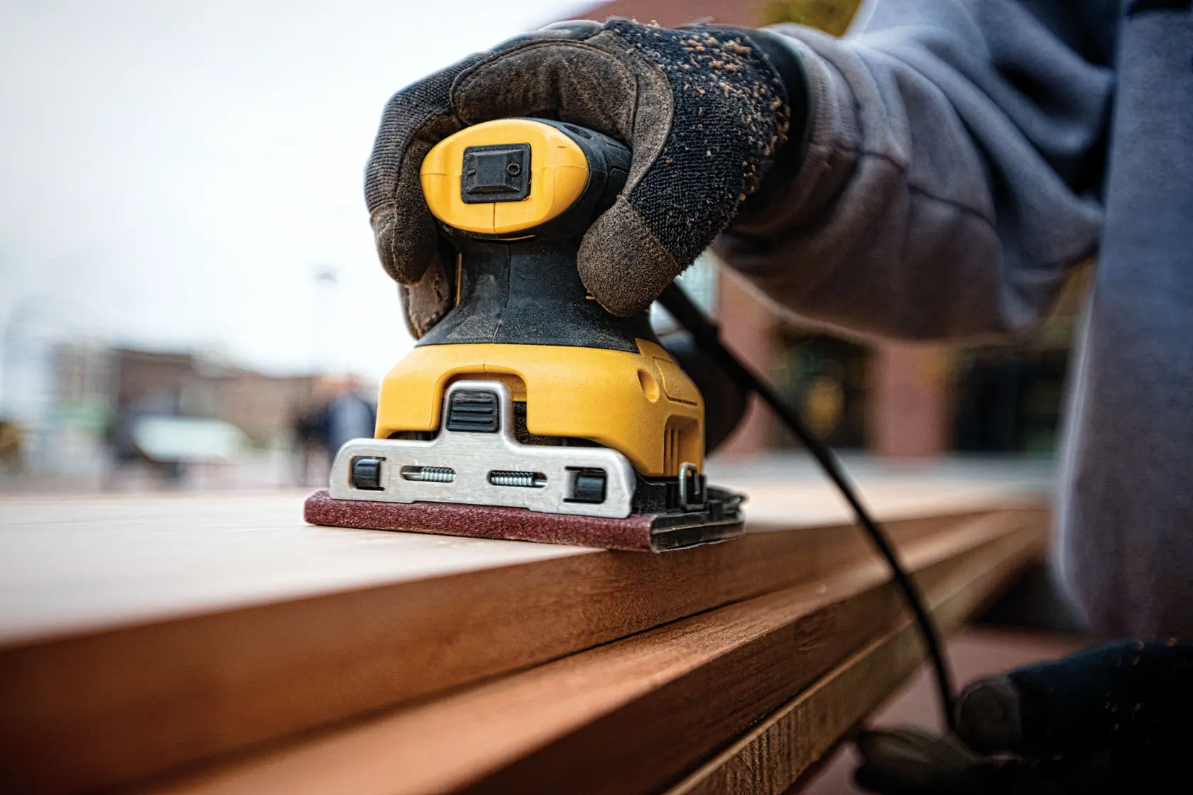 A person is using a DEWALT DWE6411 electric sander on a wooden surface inside a workshop, with the sander held vertically and dust bag attached.