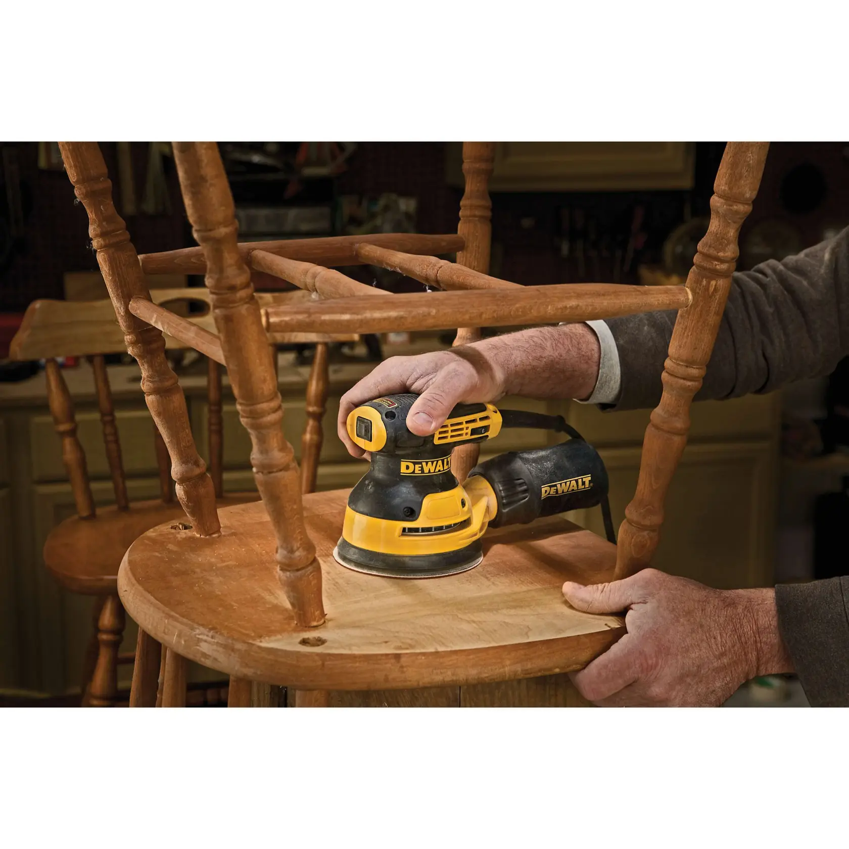 A person using a DEWALT DWE6423 electric sander to smooth the surface of a wooden chair in a workshop.