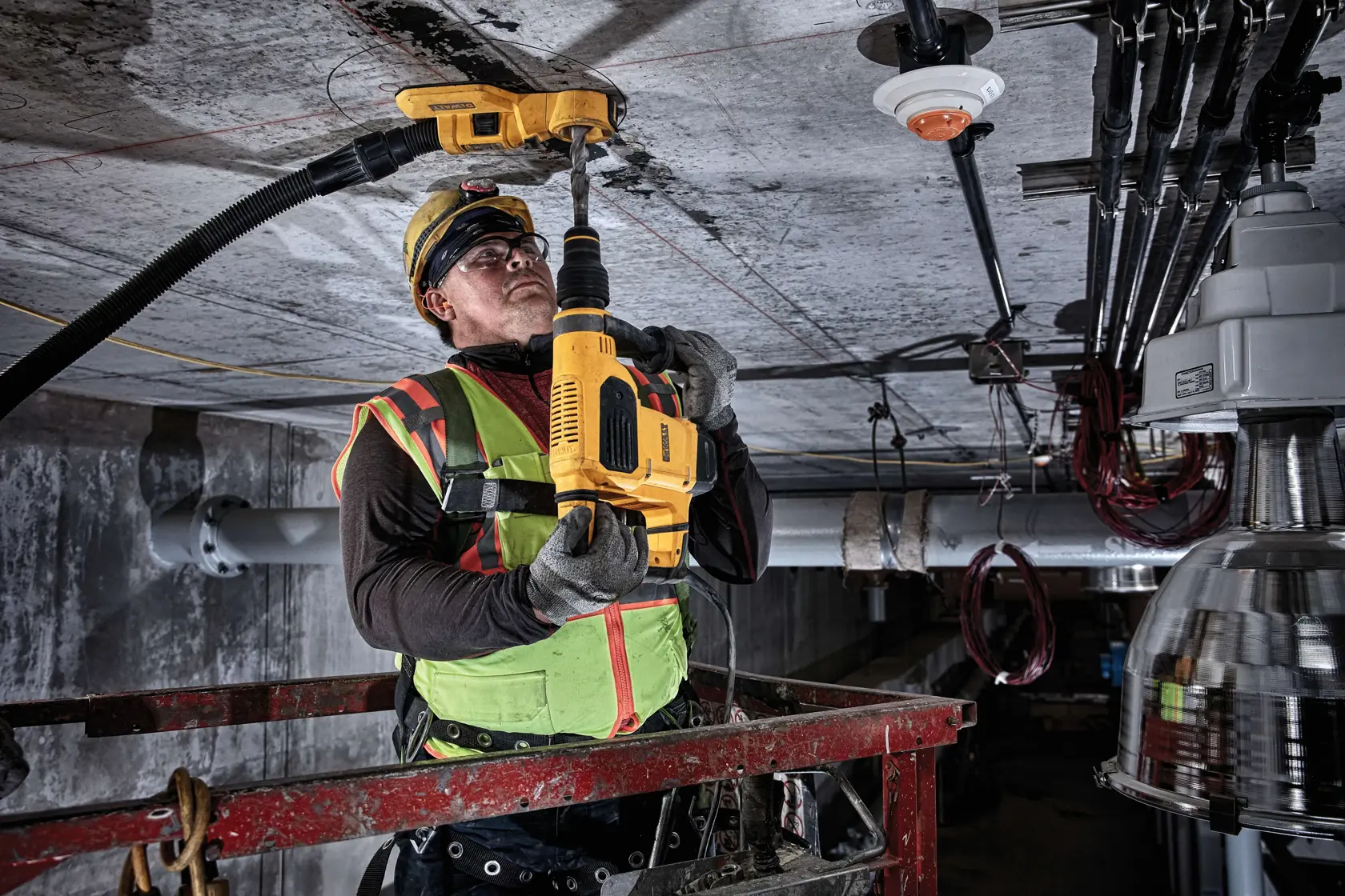 Un ouvrier du bâtiment équipé de vêtements de sécurité utilise un outil DEWALT DWH050 pour percer un plafond dans un environnement industriel. L’ouvrier se tient sur une plateforme élévatrice et un accessoire d’aspiration de poussière est raccordé à l’outil.