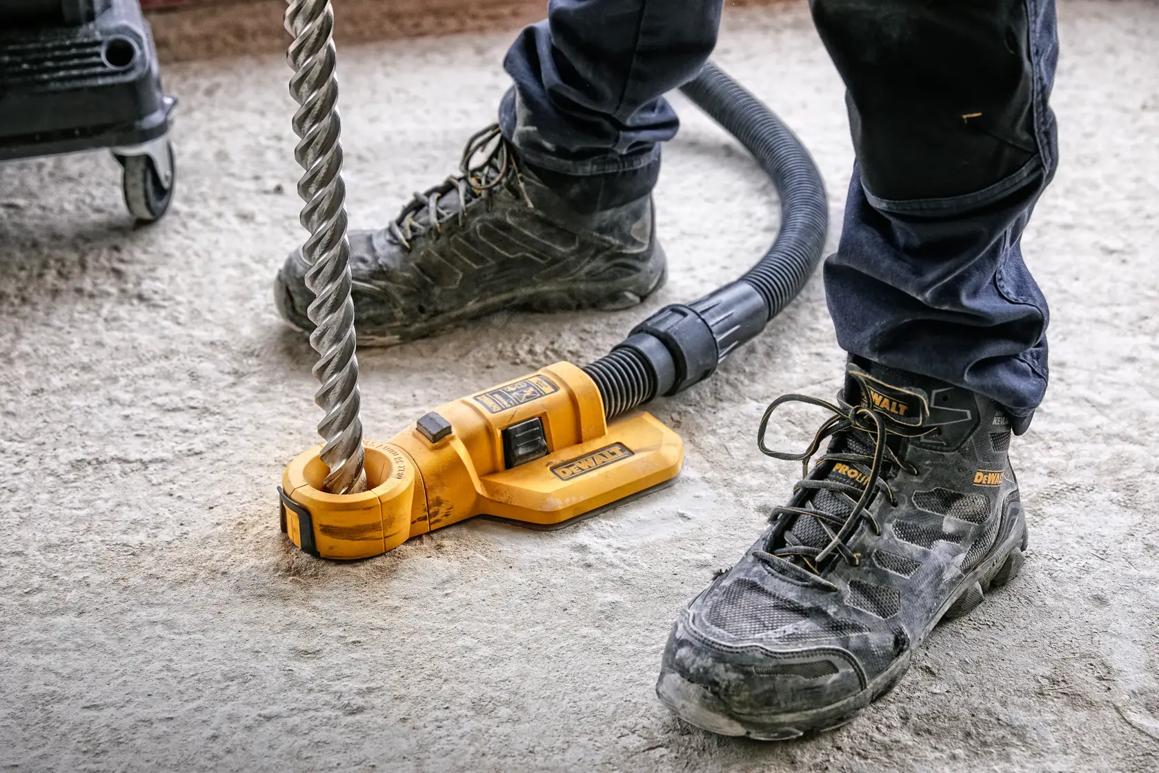 Un accessoire d'extraction de poussière DEWALT DWH050 est connecté à un tuyau d'aspirateur et positionné autour d'une grande mèche de forage sur le sol d'un chantier. Les pieds d'une personne portant des bottes de travail DEWALT et un pantalon sombre sont visibles.