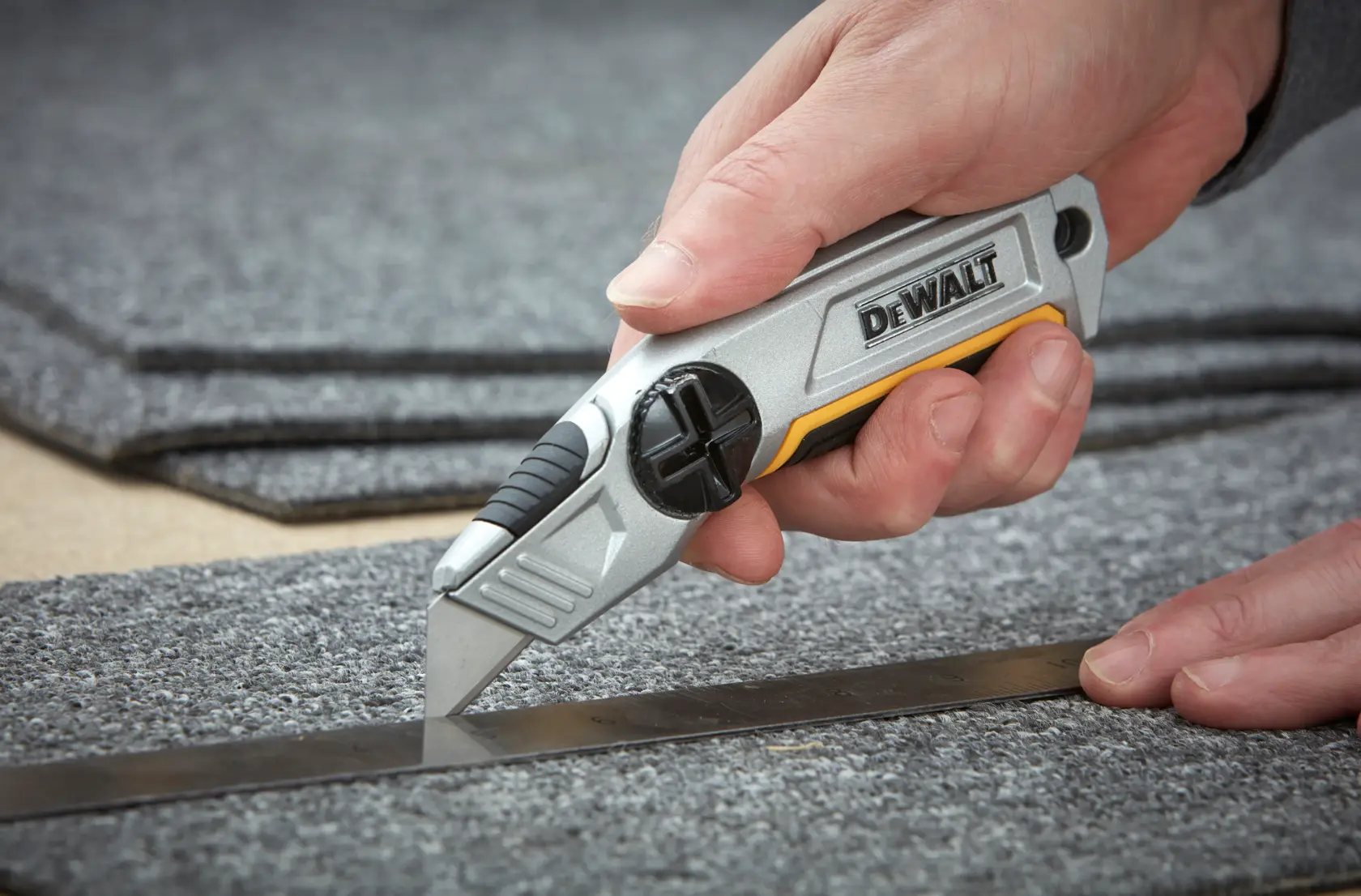 A hand uses a DEWALT utility knife to cut grey carpet along a metal ruler.