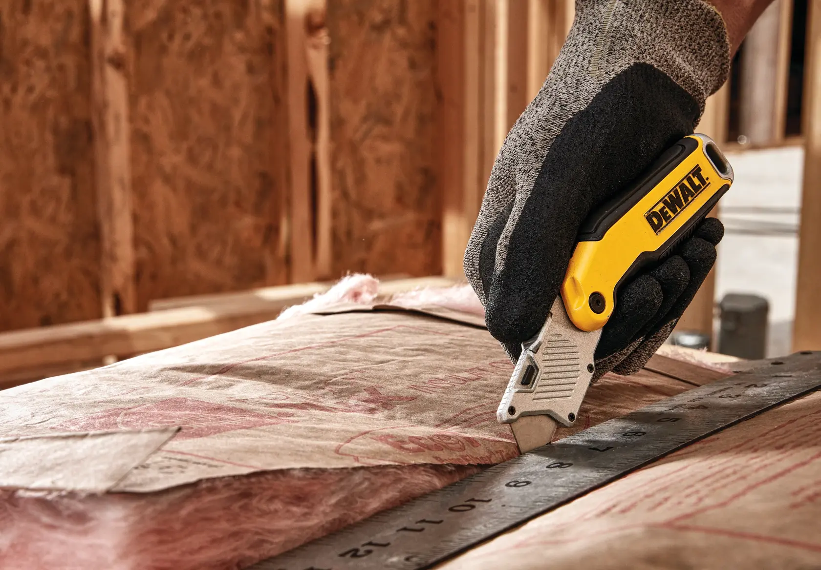 A person wearing work gloves uses a DEWALT utility knife to cut insulation material along a metal ruler in a construction setting.