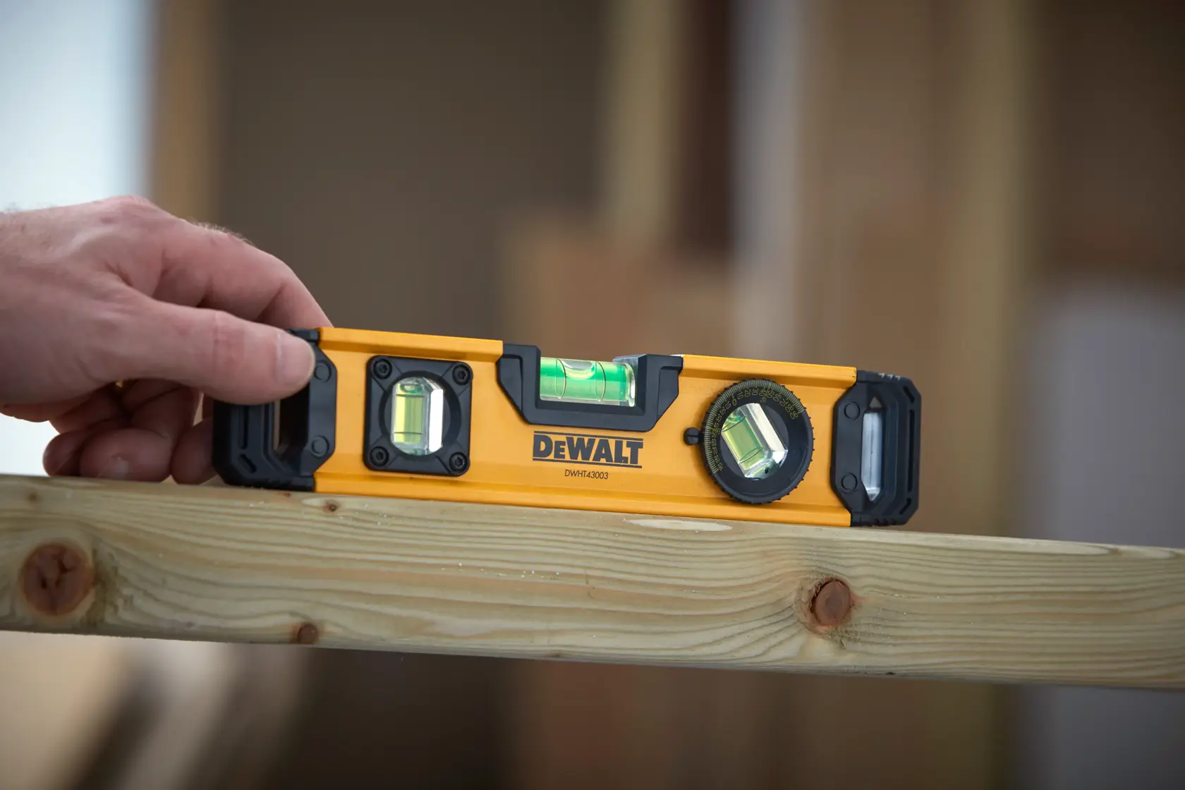 A hand is holding a DEWALT DWHT43003 spirit level tool on top of a wooden beam inside a construction area. The spirit level has three bubble vials and a yellow body.